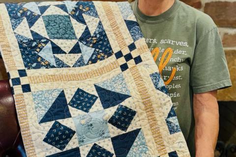 Merry White proudly displays a handcrafted quilt made by her sister, Leura (Chesser) Marler, which Marler donated to the Seminole Nation Museum’s 52nd Annual Holiday Bazaar in Wewoka. The quilt, featuring striking geometric patterns in shades of blue an