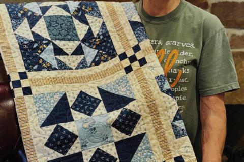 Merry White proudly displays a handcrafted quilt made by her sister, Leura (Chesser) Marler, which Marler donated to the Seminole Nation Museum’s 52nd Annual Holiday Bazaar in Wewoka. The quilt, featuring striking geometric patterns in shades of blue an